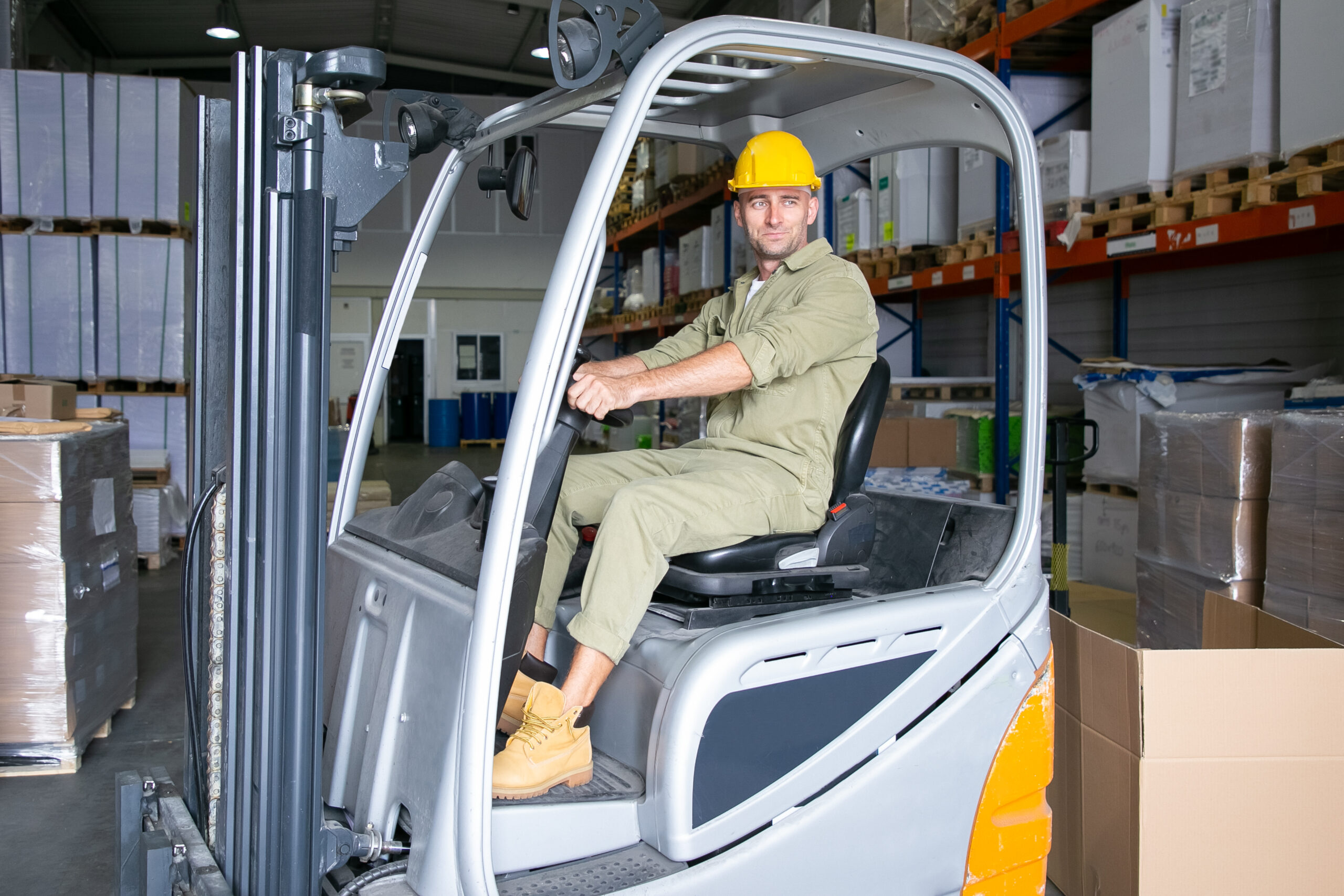Positive male logistic worker in hardhat driving forklift in warehouse, smiling, looking away. Shelves with goods on wooden trays in background. Blue collar or labor concept